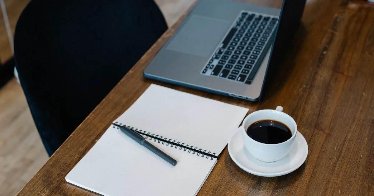 Laptop and spiral notebook with pen and coffee on a desk, representing the best note taking app for students alongside handwritten notes