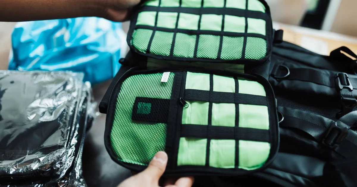 An open elastic-webbing electronics organizer held over a backpack, where a cable organizer bag price is decided by the zipper and the panel
