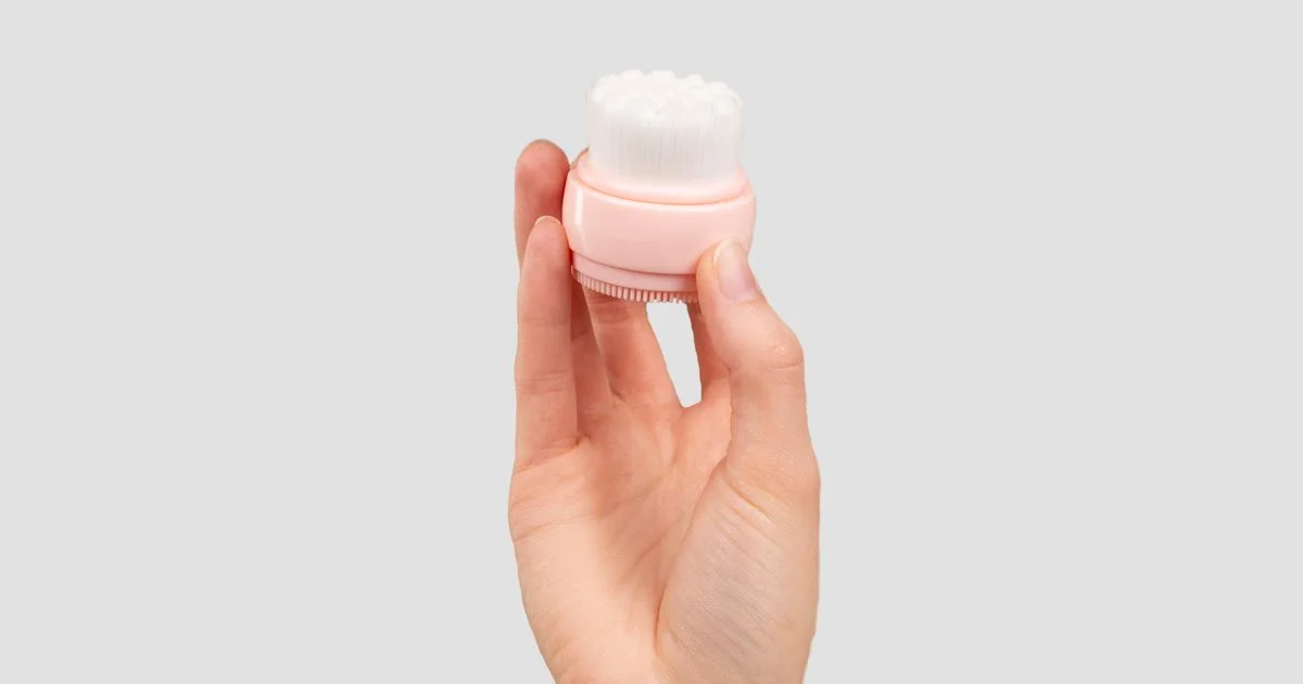 A hand holding a dual-sided cleansing brush with bristles and silicone, typical of a sonic facial cleansing brush under $60