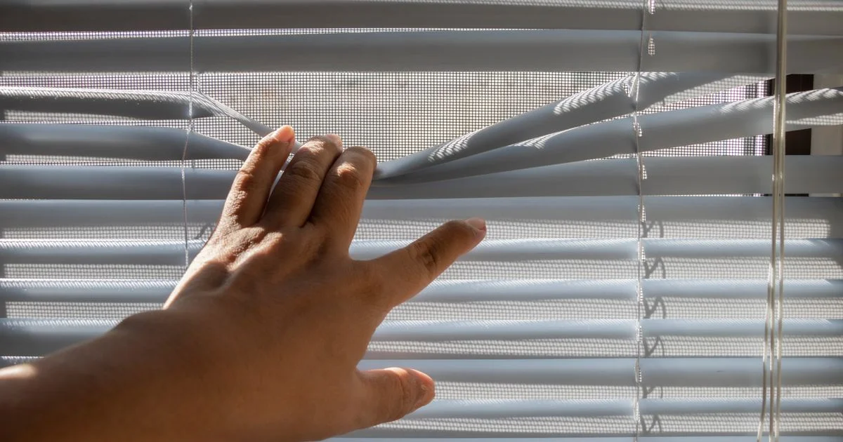 A hand parting the slats of a white venetian blind, the movement cheap smart blinds are bought to automate